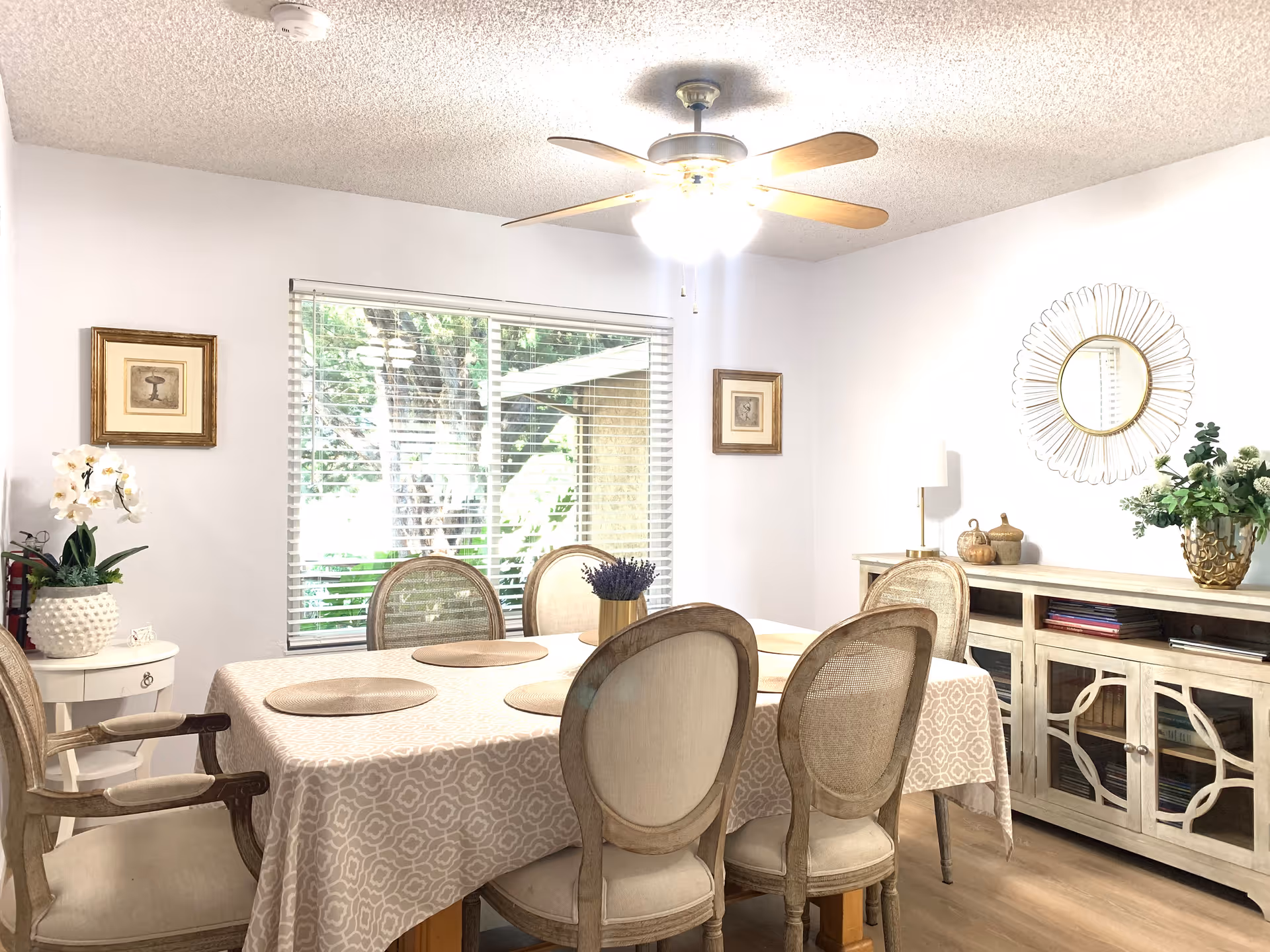 A bright dining room with a rectangular table covered by a patterned tablecloth and six cushioned chairs around it. The room features a ceiling fan with lights, a large window with blinds showing greenery outside, framed artwork on the walls, a sideboard with decorative items and books, and a small round table with a potted orchid.