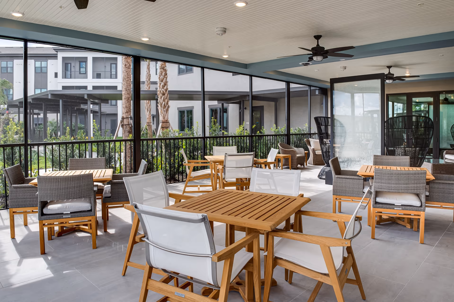 Covered outdoor patio area with multiple wooden tables and chairs, some with white cushions and others with gray woven seats. The patio is enclosed with black metal railings and has ceiling fans. In the background, there are plants, palm trees, and a view of the building exterior.