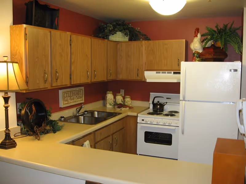 A compact kitchen with wooden cabinets, a white refrigerator, a white stove with a kettle on it, a double sink, and a beige countertop. The walls are painted red, and there are decorative items including a lamp, a rooster figurine, plants, and a framed sign that says 'Celebrate Family, Friends, & Laughter'.