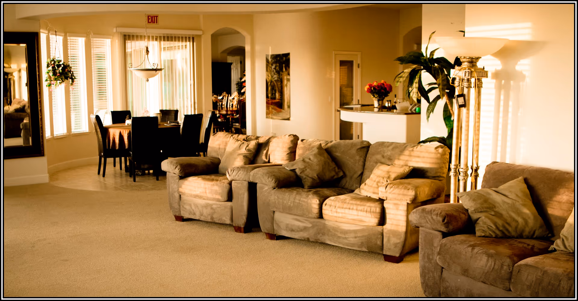 A cozy living room area with three brown cushioned sofas arranged in a row. There is a tall floor lamp with a decorative base and a potted plant next to it. In the background, a dining area with a round table covered with a brown tablecloth and six black chairs is visible. Large windows with vertical blinds allow natural light to fill the space, and a large mirror is mounted on the left wall.