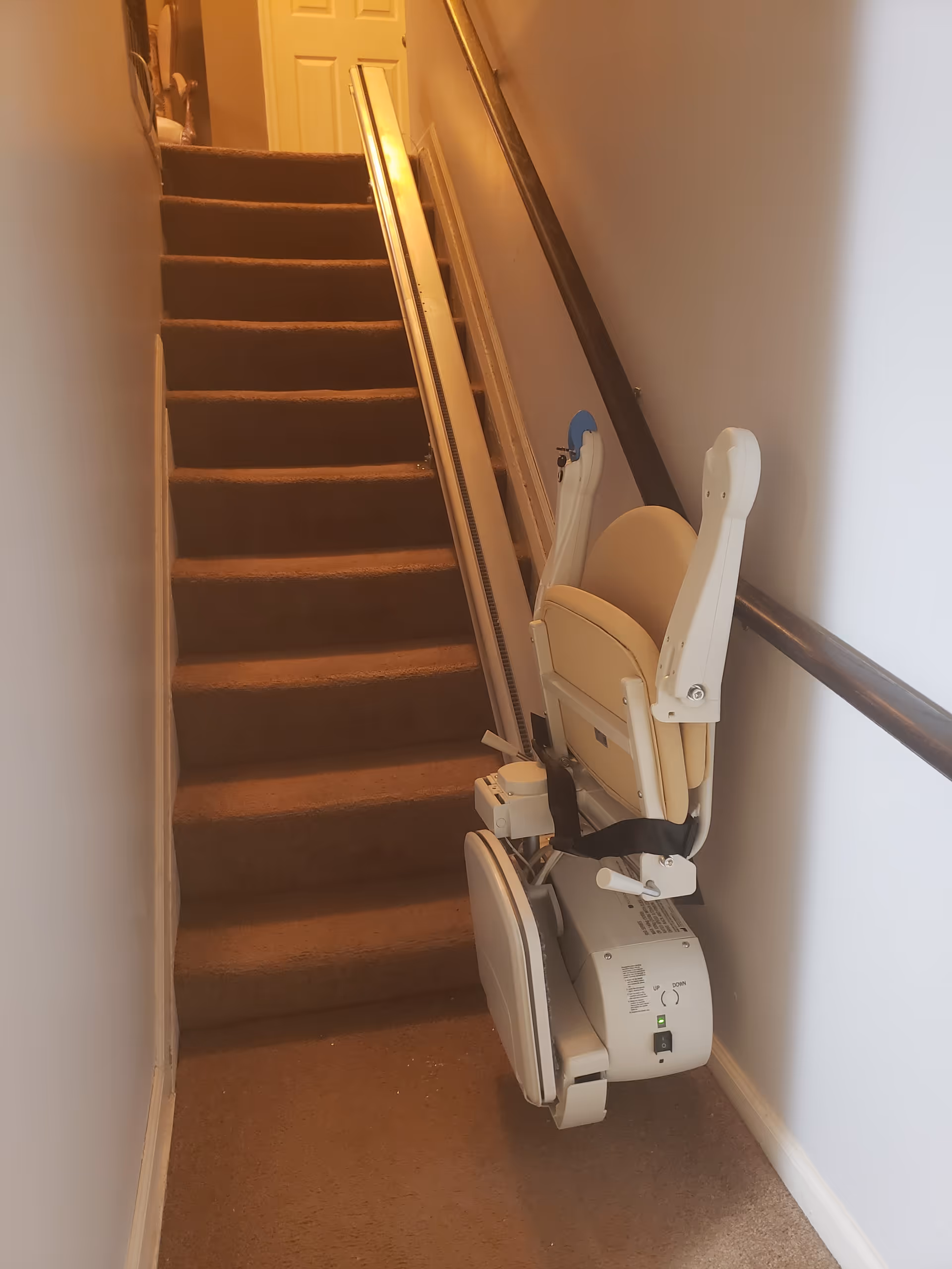 A carpeted narrow staircase with a beige stairlift installed along the right-hand wall leading up to a closed door.