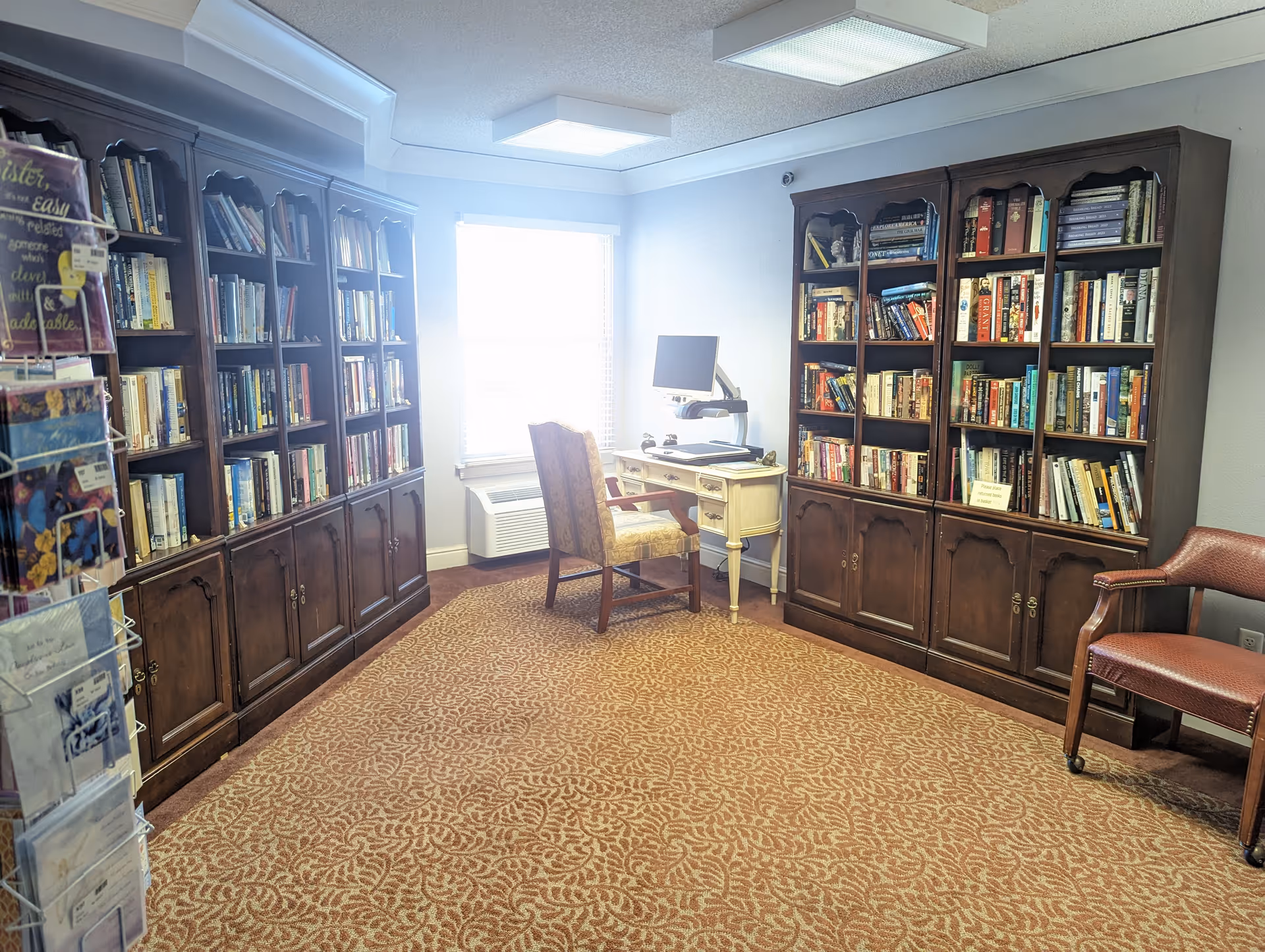 A cozy reading room with two large wooden bookshelves filled with books, a window with blinds letting in natural light, a small white desk with a computer monitor on an adjustable arm, an upholstered chair in front of the desk, and another wooden chair with red upholstery on wheels. A carpet with a patterned design covers the floor, and a rack with greeting cards is partially visible on the left side.