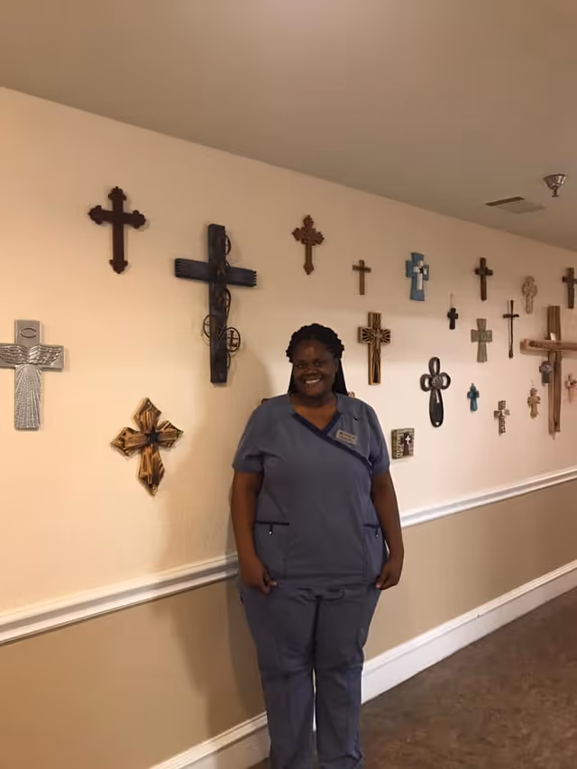 A smiling woman in blue scrubs stands in a hallway with a beige wall decorated with various styles and sizes of crosses.