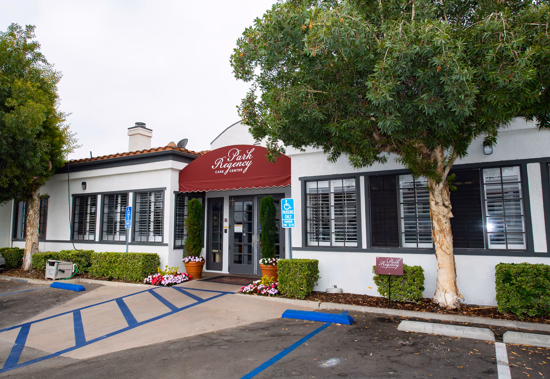 Front entrance of Park Regency Care Center with a red awning, double doors, handicap parking spaces and surrounding trees and flowers.