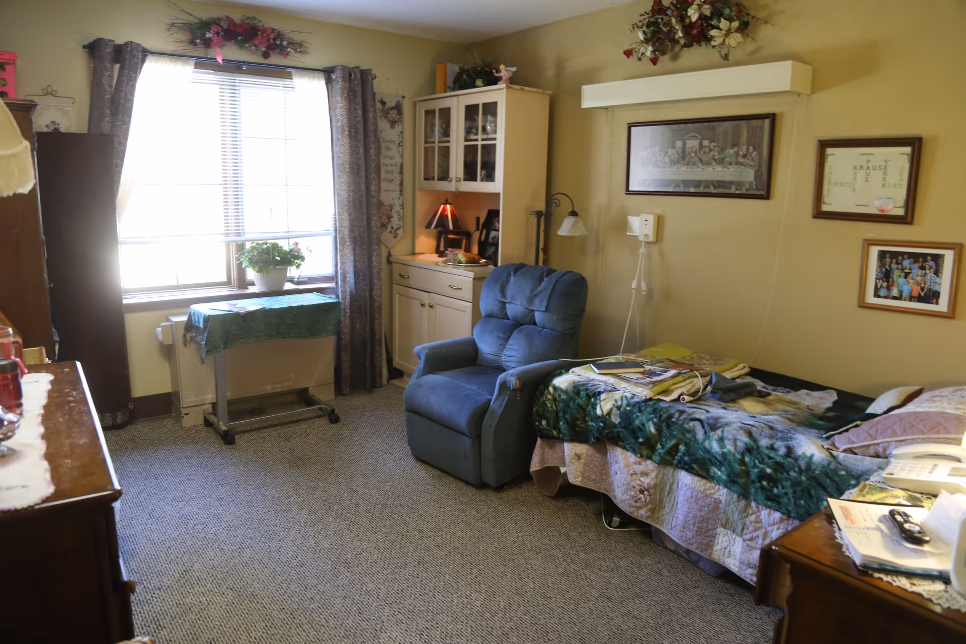 A cozy bedroom in a senior living facility with a single bed covered with a green and beige blanket, a blue recliner chair, a small cabinet with a lamp, and a window with curtains letting in natural light. The walls are decorated with framed pictures and floral arrangements.