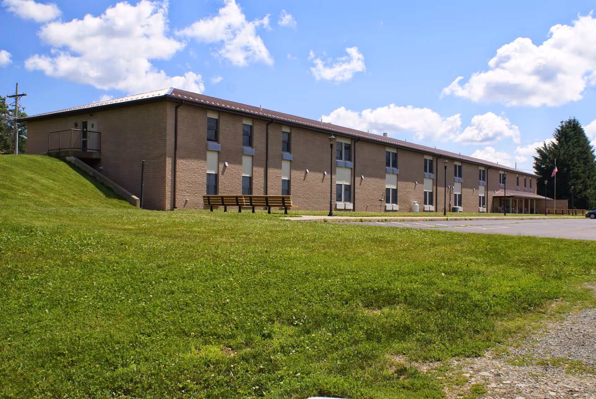 Long two-story brick building with a large grassy lawn, benches, and a parking area under a blue sky.