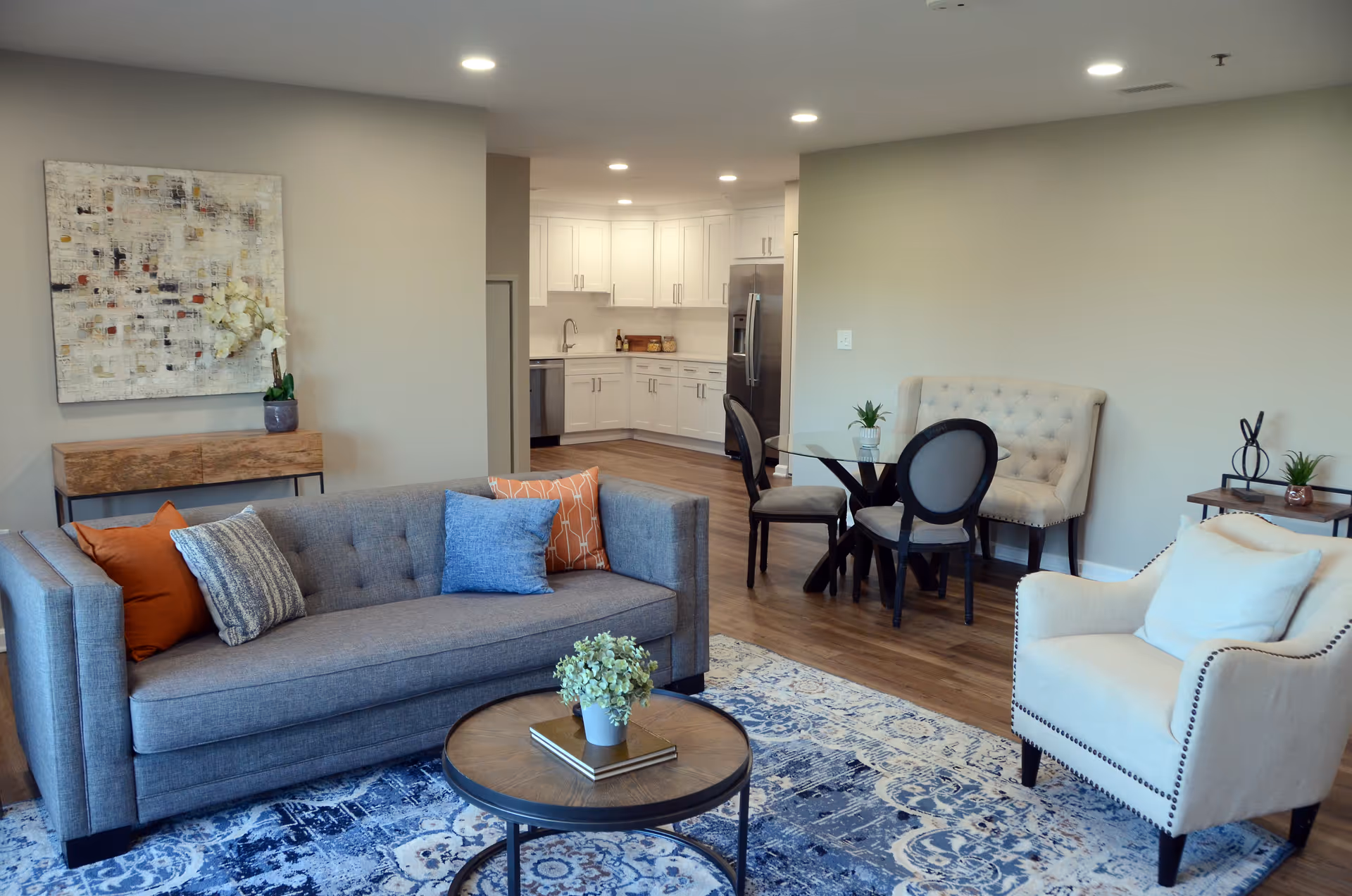 A modern living room with a gray sofa adorned with orange, blue, and patterned pillows, a round wooden coffee table with a small plant and books, a white armchair with a cushion, and a blue patterned rug. In the background, there is a small dining area with a glass-top table, two chairs, and a tufted bench. The kitchen with white cabinets, stainless steel appliances, and wooden flooring is visible beyond the dining area. A piece of abstract art and a small wooden console table with a plant are on the left wall.