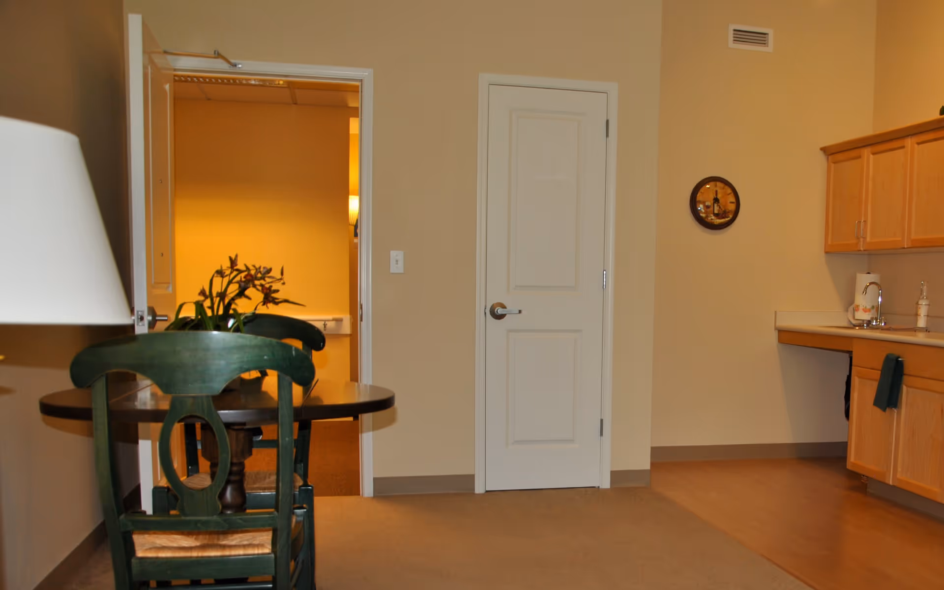 Interior view of a room with beige walls and carpeted floor. There is a small round wooden table with two green chairs, a lamp on the left side, and a potted plant on the table. To the right, there is a kitchenette area with wooden cabinets, a countertop, a sink, and a towel hanging from the cabinet. Two doors are visible on the far wall, one open showing a hallway with a yellowish light and the other closed. A decorative clock hangs on the wall above the kitchenette.