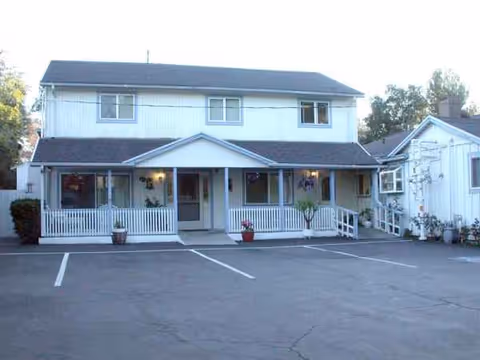 Two-story light blue building with a front porch and potted plants in a parking lot.
