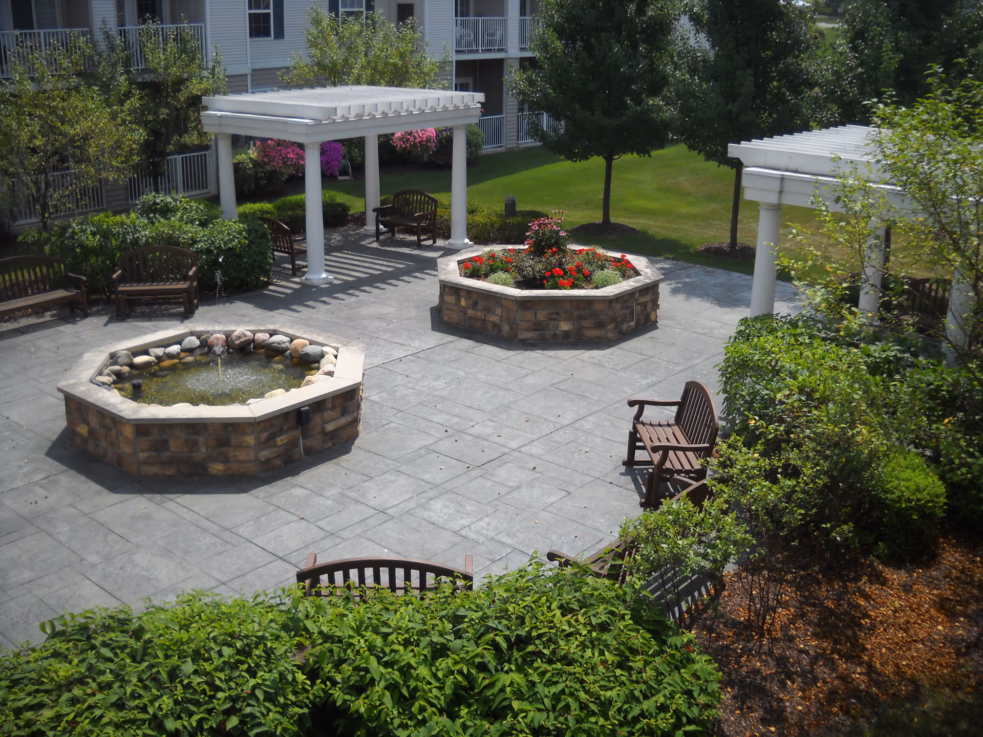 Outdoor courtyard area with stone-paved ground, featuring two octagonal stone planters, one with a small water fountain and the other with red flowers. Several wooden benches are placed around the courtyard, along with white pergolas providing partial shade. Surrounding the courtyard are green bushes, trees, and a multi-story building with balconies in the background.