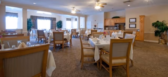 A spacious dining room with multiple tables covered in white tablecloths, each set with napkins, glasses, and silverware. Wooden chairs surround the tables. The room has beige walls, carpeted floors, ceiling fans with lights, and large windows with curtains allowing natural light. There are plants and framed pictures on the walls, and a wooden cabinet area in the back.