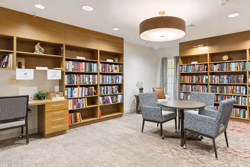 A cozy library room with wooden bookshelves filled with books along the walls. There is a round table with four upholstered chairs in the center of the room. A desk with a chair is positioned against one wall, and the room is well-lit with ceiling lights and a large circular pendant light. The carpet has a subtle floral pattern, and a window with curtains is visible in the background.