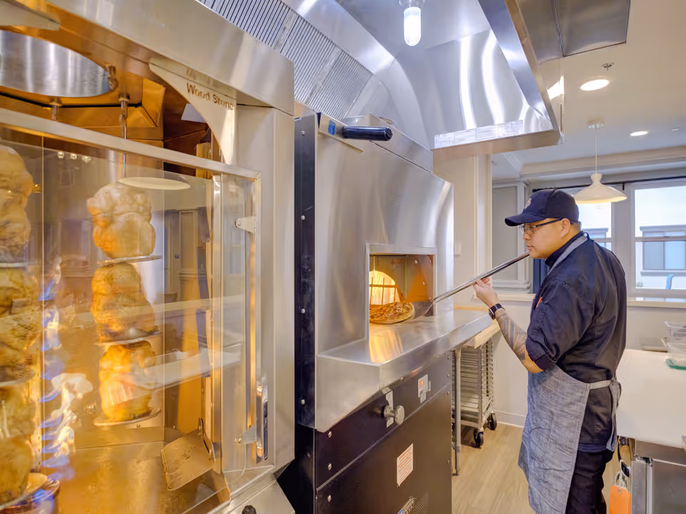 A cook uses a pizza peel to remove a pizza from a stainless steel wood-fired oven in a commercial kitchen with a rotisserie visible on the left.