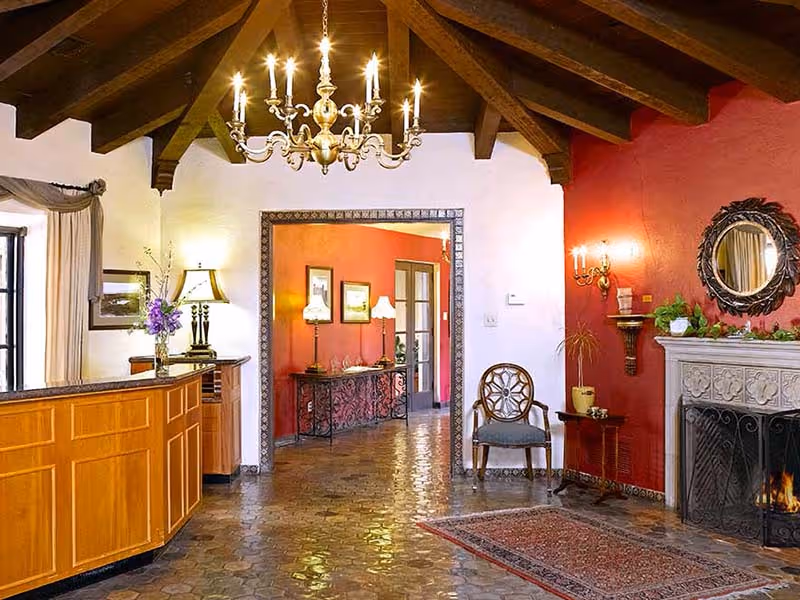 A warmly lit interior space with a wooden reception desk on the left, a chandelier hanging from a wooden beamed ceiling, a stone fireplace with a fire burning on the right, a decorative mirror above the fireplace, a chair and small table near the fireplace, and a hallway leading to another room with red walls and lamps.