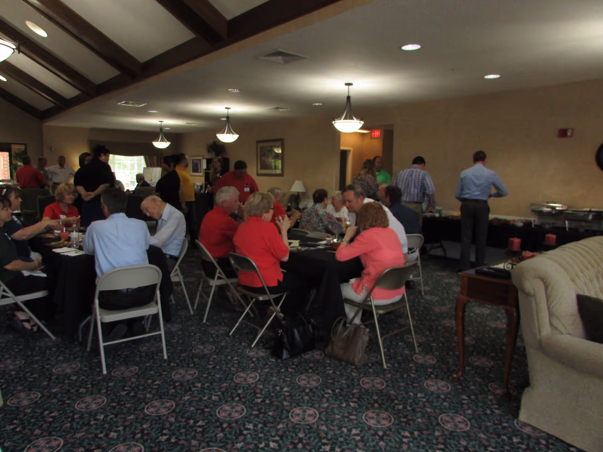 People gathered around tables in a carpeted community dining room with a buffet table along the wall.