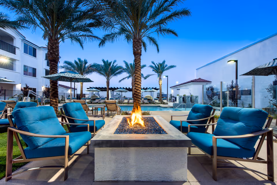 Outdoor poolside courtyard with a central fire pit surrounded by blue-cushioned lounge chairs, palm trees, and buildings in the background.