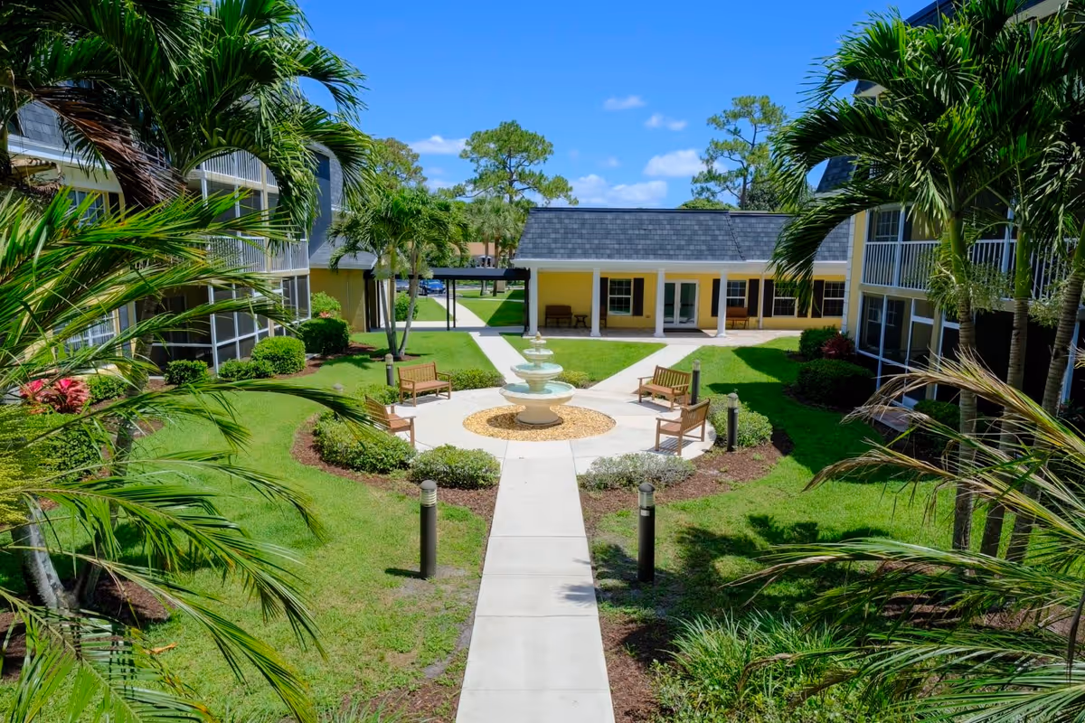 Sunlit courtyard with a central fountain, benches, palm trees and surrounding two-story buildings.