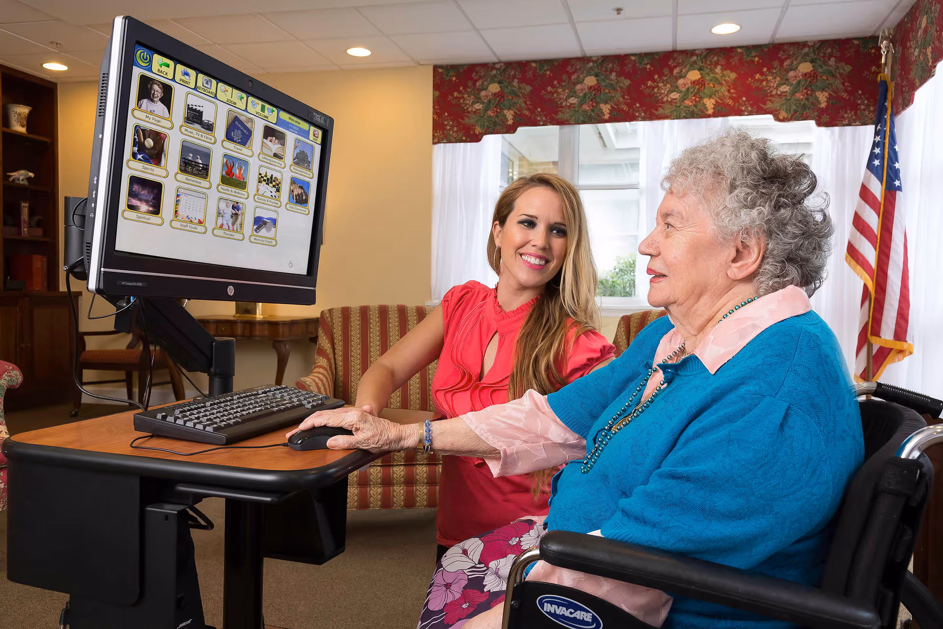 An elderly woman in a wheelchair using a computer with the assistance of a younger woman in a red blouse, both smiling. They are in a cozy room with a striped couch, a wooden table, and an American flag in the background.