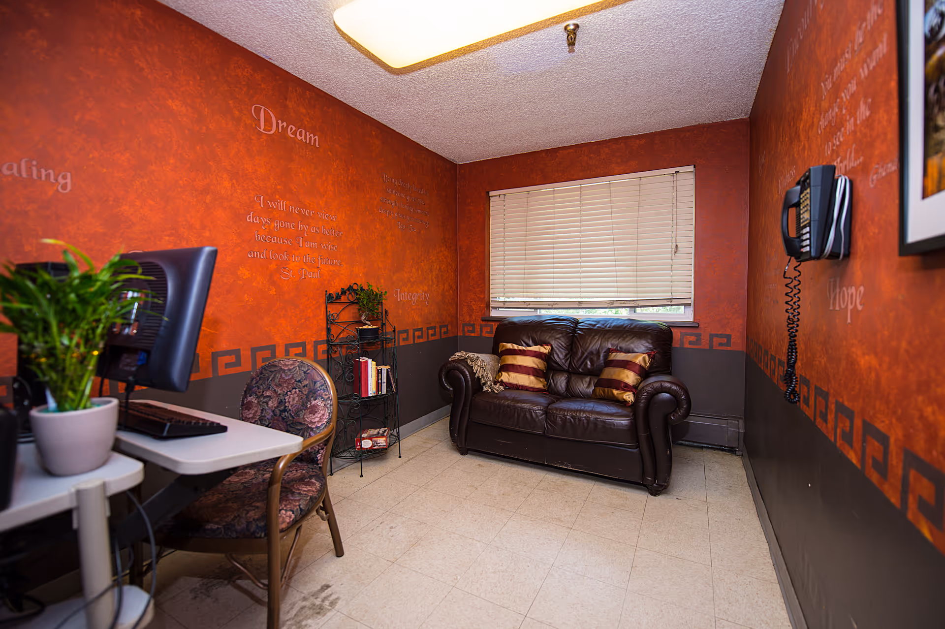 Small sitting room with a brown leather loveseat, computer desk and chair, potted plant, blinds, and red textured walls with a wall phone.