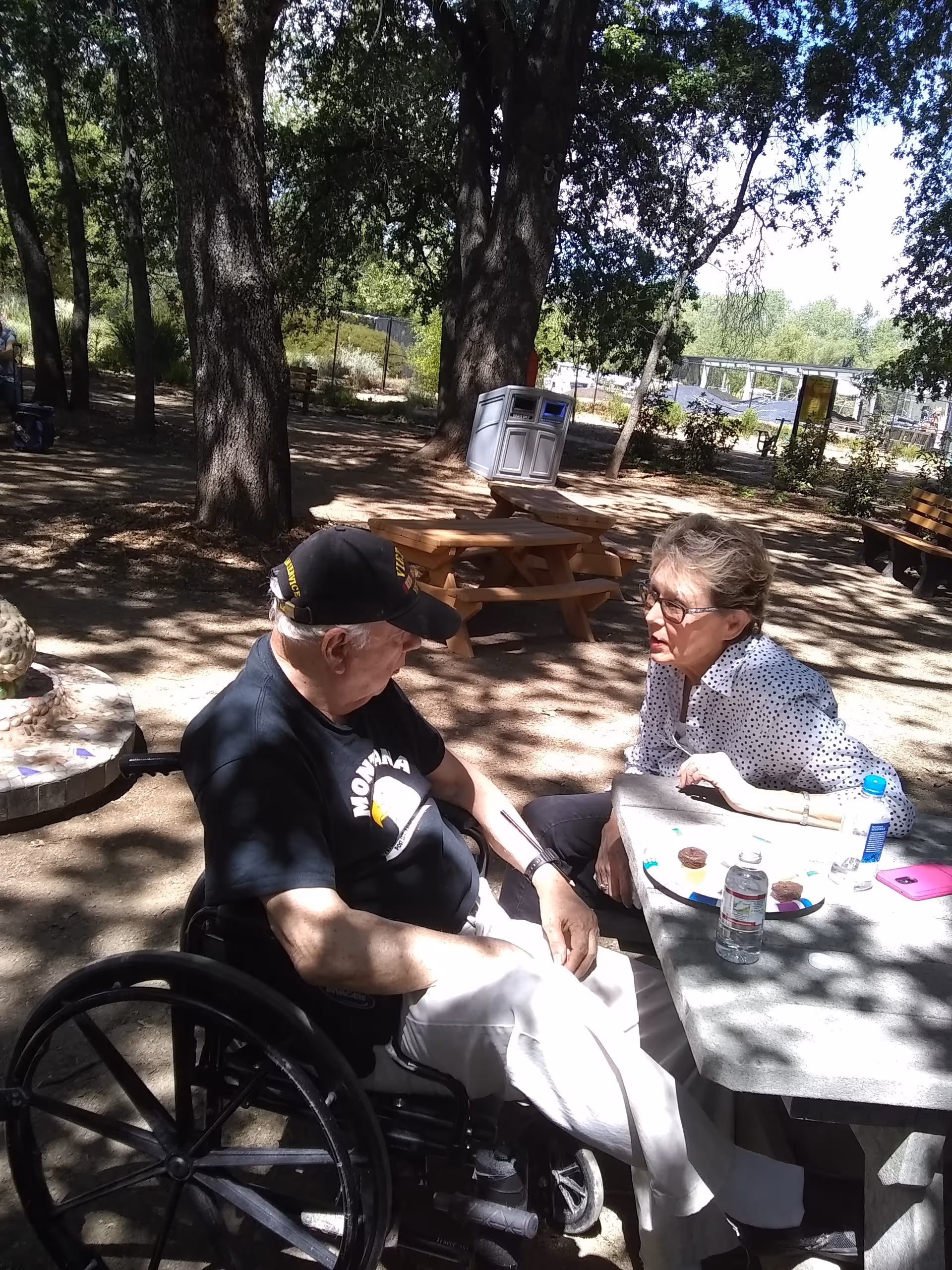 An elderly man in a wheelchair and an elderly woman sitting at a picnic table outdoors in a shaded park area with trees and benches around them. The man is wearing a black cap and black t-shirt, and the woman is wearing glasses and a white blouse with black dots. On the table are a water bottle, a plate with some food, and a pink phone.