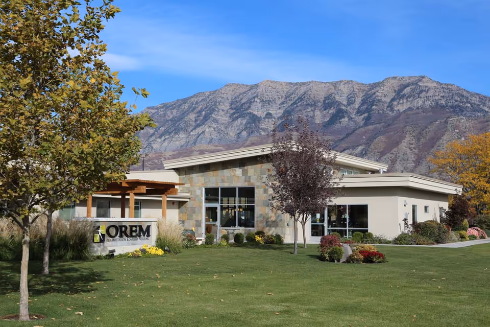 Front view of Orem Rehabilitation & Skilled Nursing single-story building with a lawn, trees, and mountains in the background.