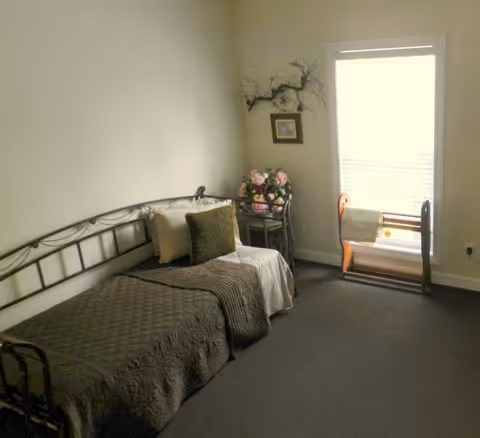 Small bedroom with a single metal-framed bed, a side table holding flowers, and a window with a wooden chair.