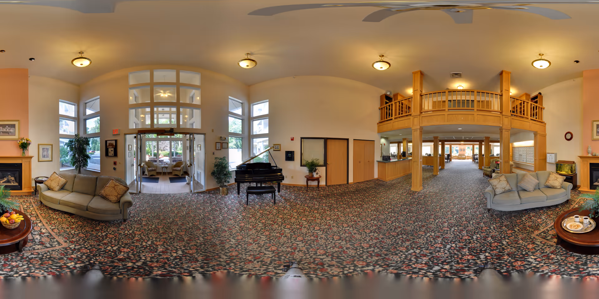 Spacious senior living facility lobby with floral carpet, two beige sofas with cushions, a grand piano, wooden pillars and balcony, large windows letting in natural light, a fireplace on each side, and a coffee table with fruit and tea set.