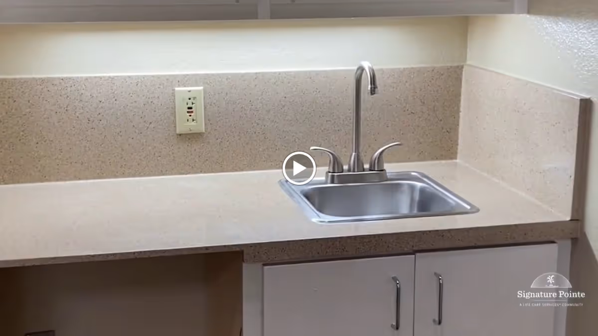 Small kitchenette countertop with a stainless steel sink and faucet, speckled backsplash and white cabinets below.