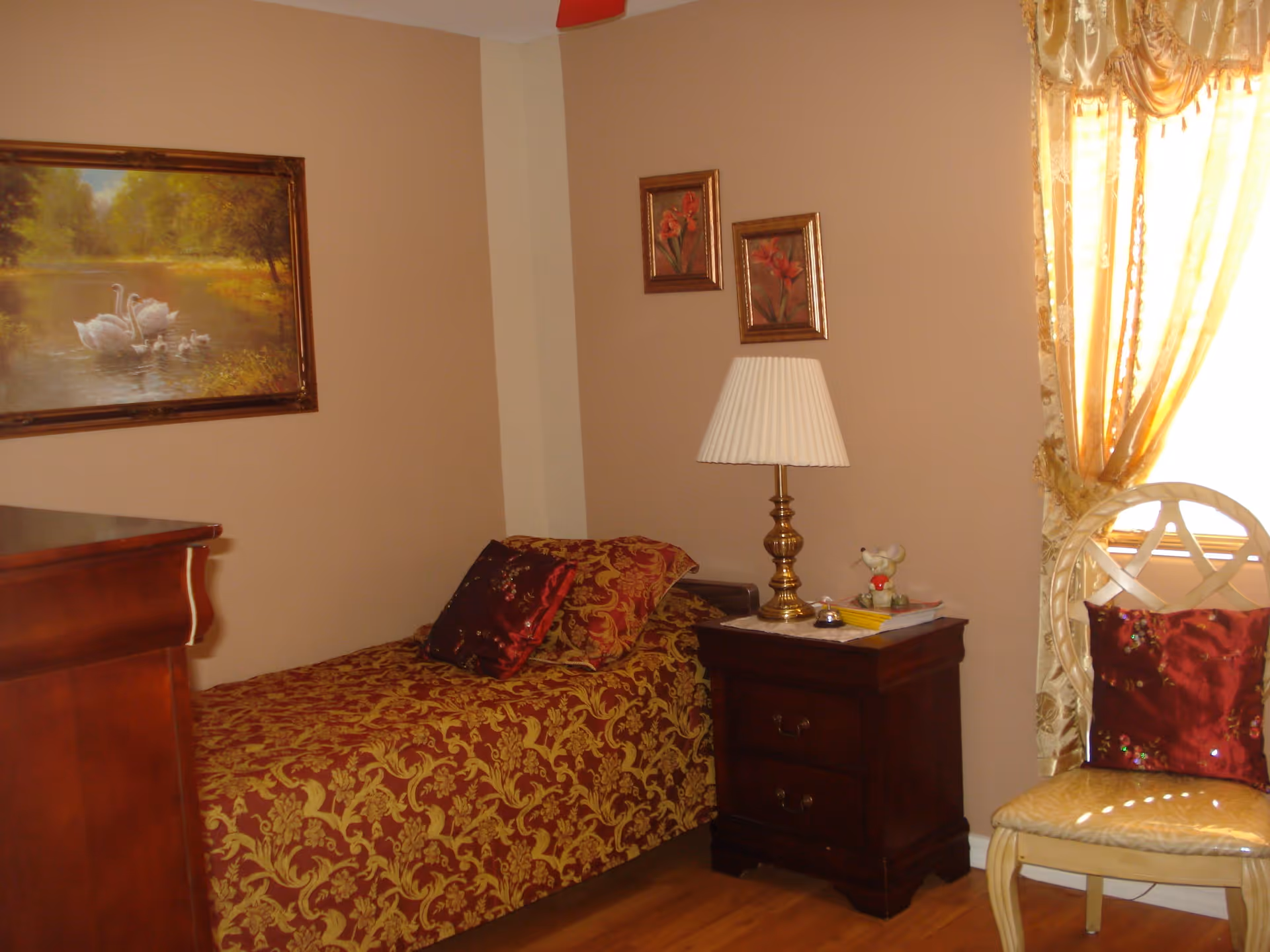A cozy bedroom with a single bed covered in a red and gold patterned bedspread and matching pillows. Next to the bed is a dark wooden nightstand with a brass lamp, a small figurine, and some books. A wooden chair with a red cushion is placed near a window with golden curtains. The walls are beige and decorated with framed paintings, including one of swans on a pond.