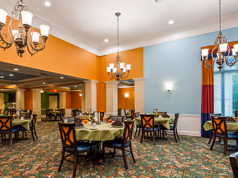 A brightly lit dining room with multiple round tables covered with green tablecloths, each set with napkins, cups, and small flower arrangements. The room features orange and light blue walls, ornate chandeliers hanging from the ceiling, and a patterned carpet. Large windows with colorful curtains allow natural light to enter.