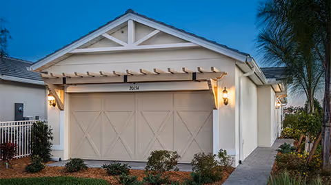 Exterior view of a single-story residential building with a closed double garage door, beige walls, and a small pergola above the garage. The house number 2034 is visible above the garage door. There are outdoor wall lanterns on either side of the garage, a paved walkway on the right, and landscaping with bushes and palm trees in front.