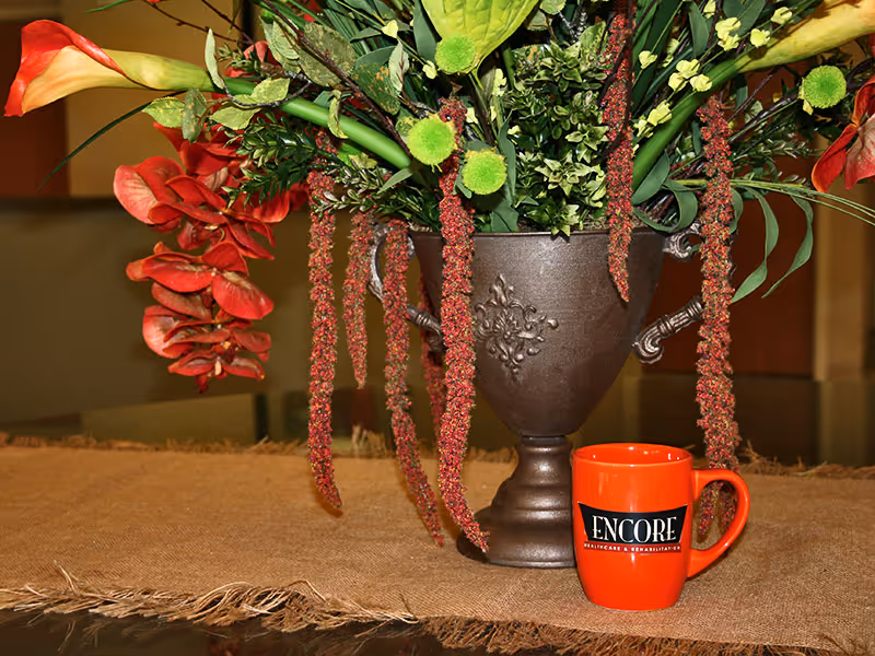 A decorative floral arrangement in a dark ornate vase with various green leaves and red flowers, placed on a burlap cloth. Next to the vase is an orange mug with the text 'ENCORE Healthcare & Rehabilitation' printed on it.