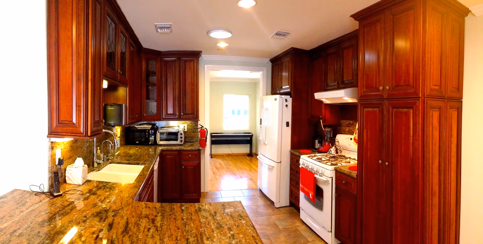 A well-lit kitchen with dark wooden cabinets, granite countertops, a white sink, a white refrigerator, a white gas stove with a red towel hanging on the oven door, and small appliances like a toaster oven and microwave. The kitchen opens into another room with wooden flooring and a window letting in natural light.