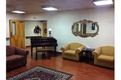 Community sitting area with upholstered chairs and sofa, a piano, decorative mirror, side table, and area rug on wood flooring.
