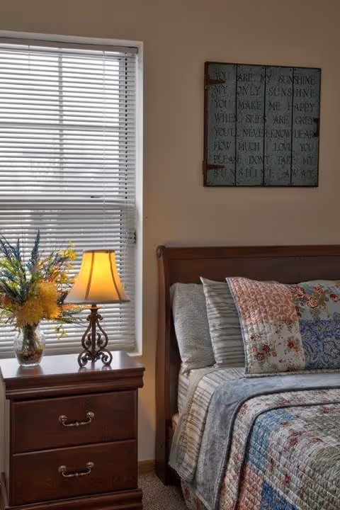 A cozy bedroom corner featuring a wooden bed with a patchwork quilt and multiple pillows. Next to the bed is a wooden nightstand with two drawers, a decorative table lamp with a yellow shade, and a vase of colorful flowers. A window with closed blinds is behind the nightstand, and a decorative wall hanging with text is above the bed.