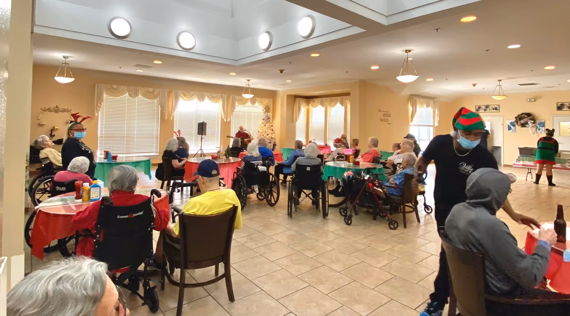 A group of elderly people seated at round tables covered with red, green, and pink tablecloths in a large, well-lit room. Some individuals are in wheelchairs, and a few staff members wearing festive holiday hats and masks are attending to them. A person is playing guitar near a white Christmas tree decorated with lights. The room has large windows with blinds and beige walls with light fixtures on the ceiling.