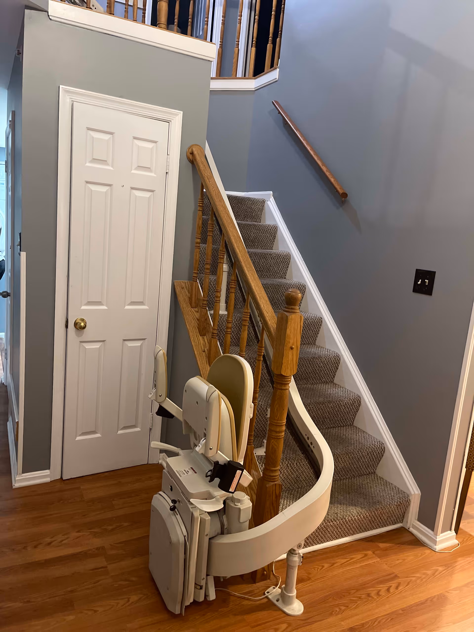 Indoor staircase with a beige stairlift installed along the wooden handrail. The stairs are carpeted in a gray pattern, and the walls are painted gray. There is a white door to the left of the staircase and a wooden floor at the base of the stairs.