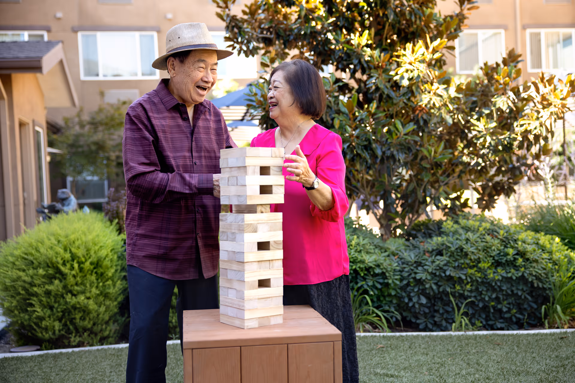 An elderly man wearing a hat and a purple shirt and an elderly woman in a bright pink blouse are playing a large outdoor Jenga game on a wooden table in a garden area with bushes and trees, smiling and enjoying their time together.