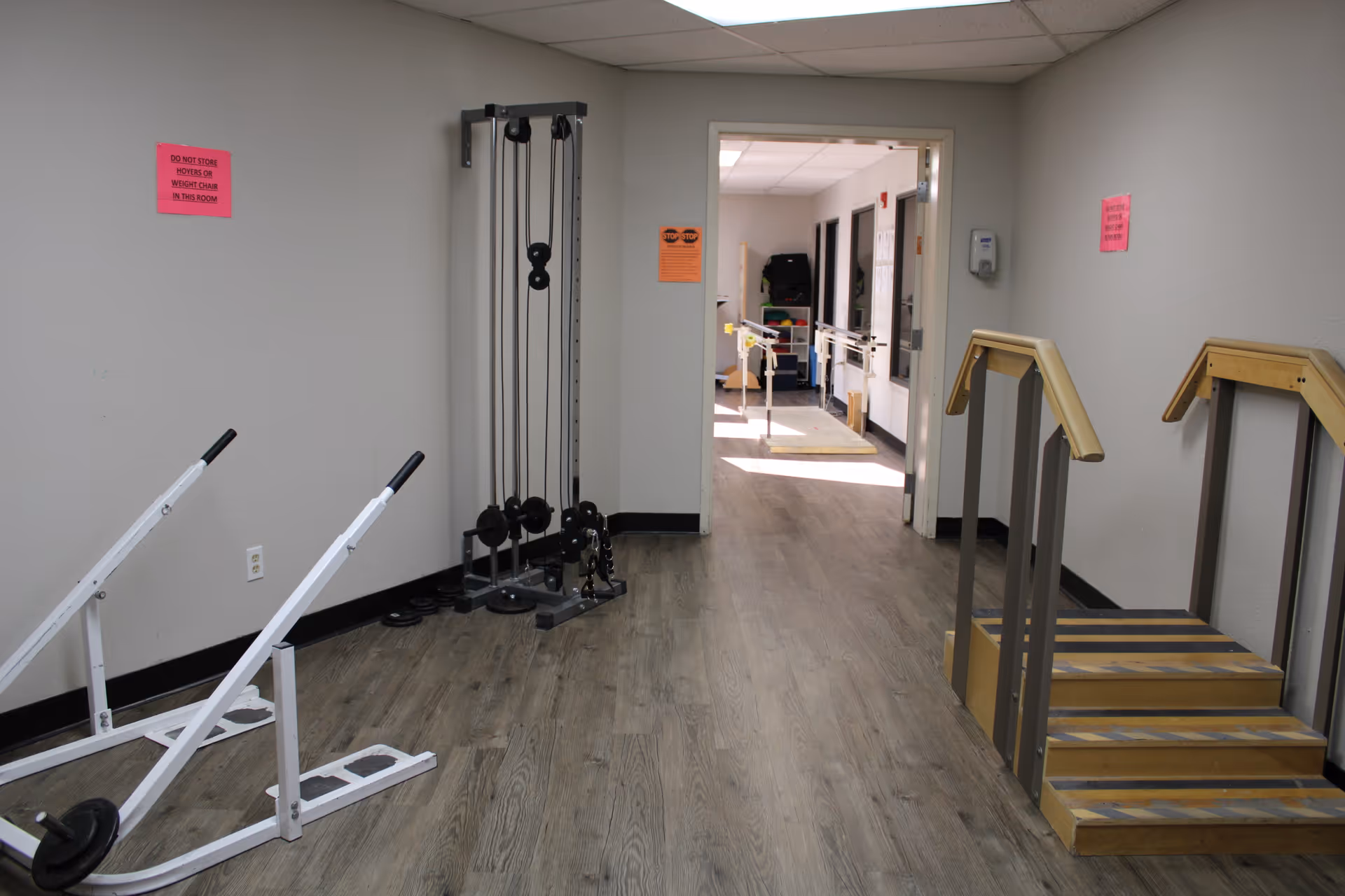 A rehabilitation room with exercise equipment including a pulley weight machine, two white sled-like exercise devices with weights, and a wooden staircase with handrails for physical therapy. The room has light-colored walls and wood-patterned flooring. There are pink and orange signs on the walls, and an open doorway leads to another room with parallel bars for walking exercises.