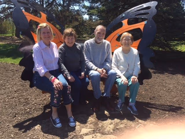 Four elderly people sitting on a large butterfly-shaped bench outdoors with trees and grass in the background, enjoying a sunny day.