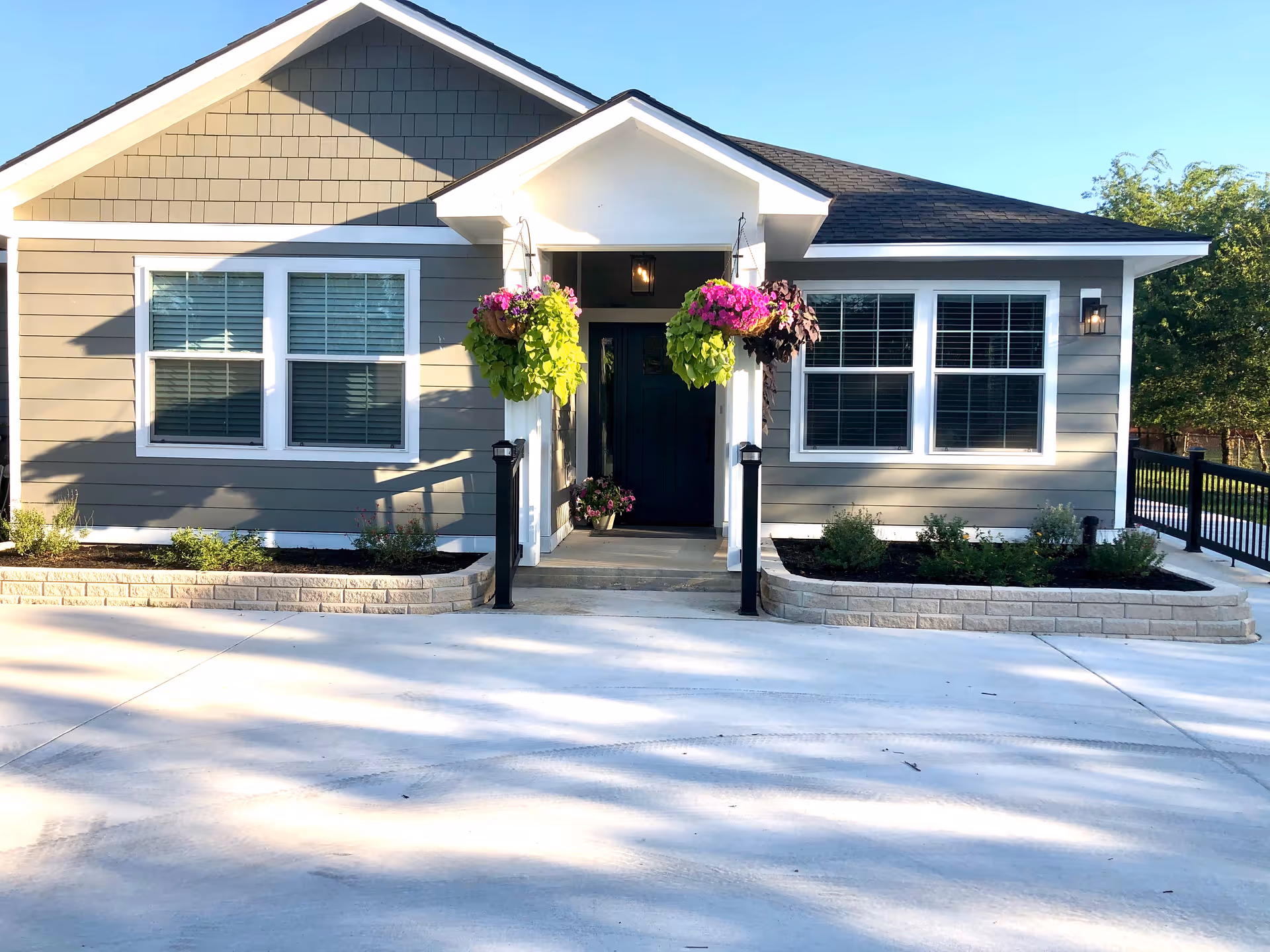 Front exterior view of a single-story senior care facility building with gray siding, white trim, and a black front door. There are two hanging flower baskets with pink and green plants flanking the entrance, and small landscaped garden beds on either side of the walkway leading to the door. The driveway and walkway are made of concrete, and there is a black metal railing on the right side.