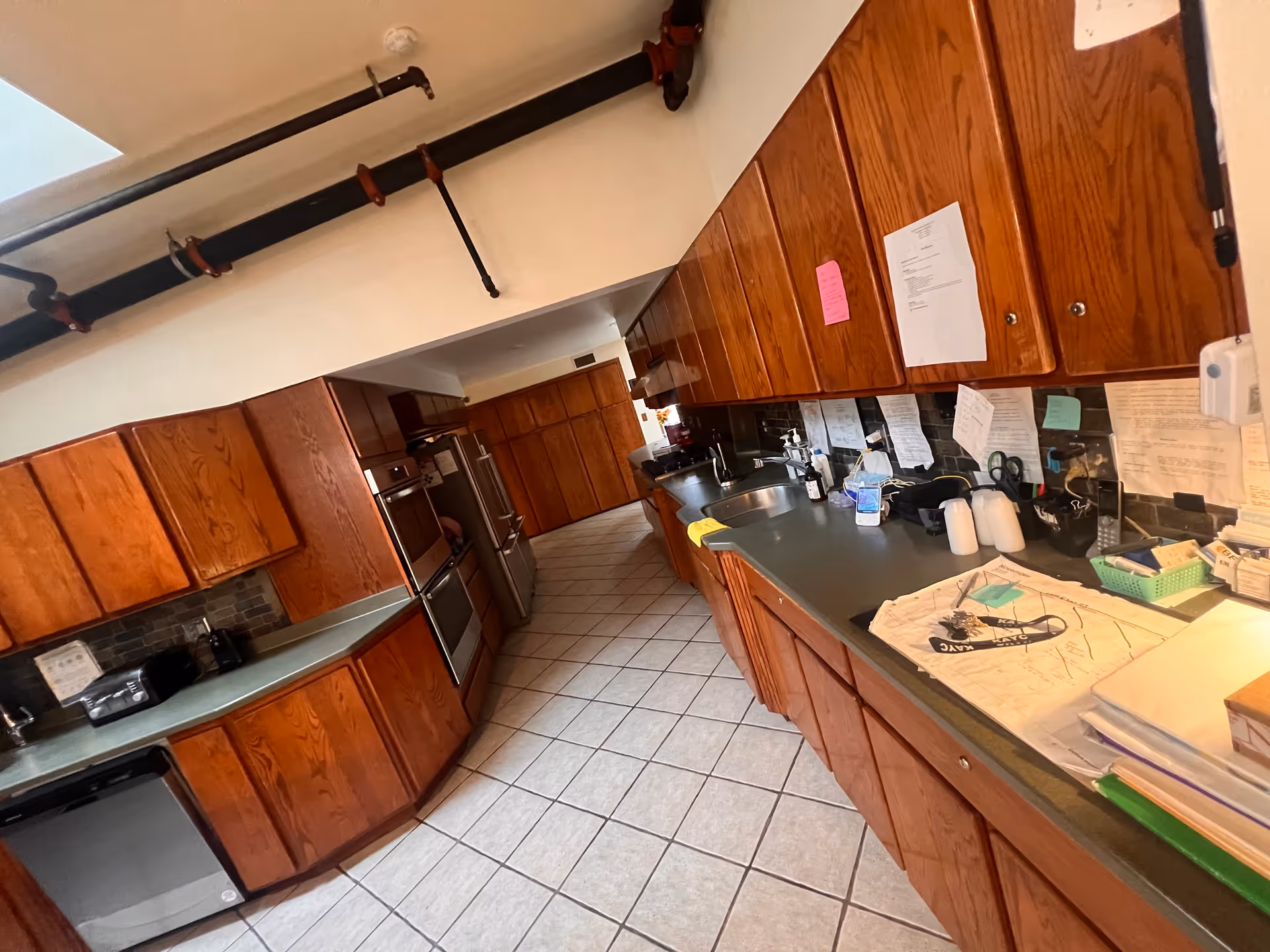 A long kitchen with wooden cabinets on both sides, a tiled floor, and various items on the countertops including papers, bottles, and kitchen appliances. There are exposed pipes on the ceiling.
