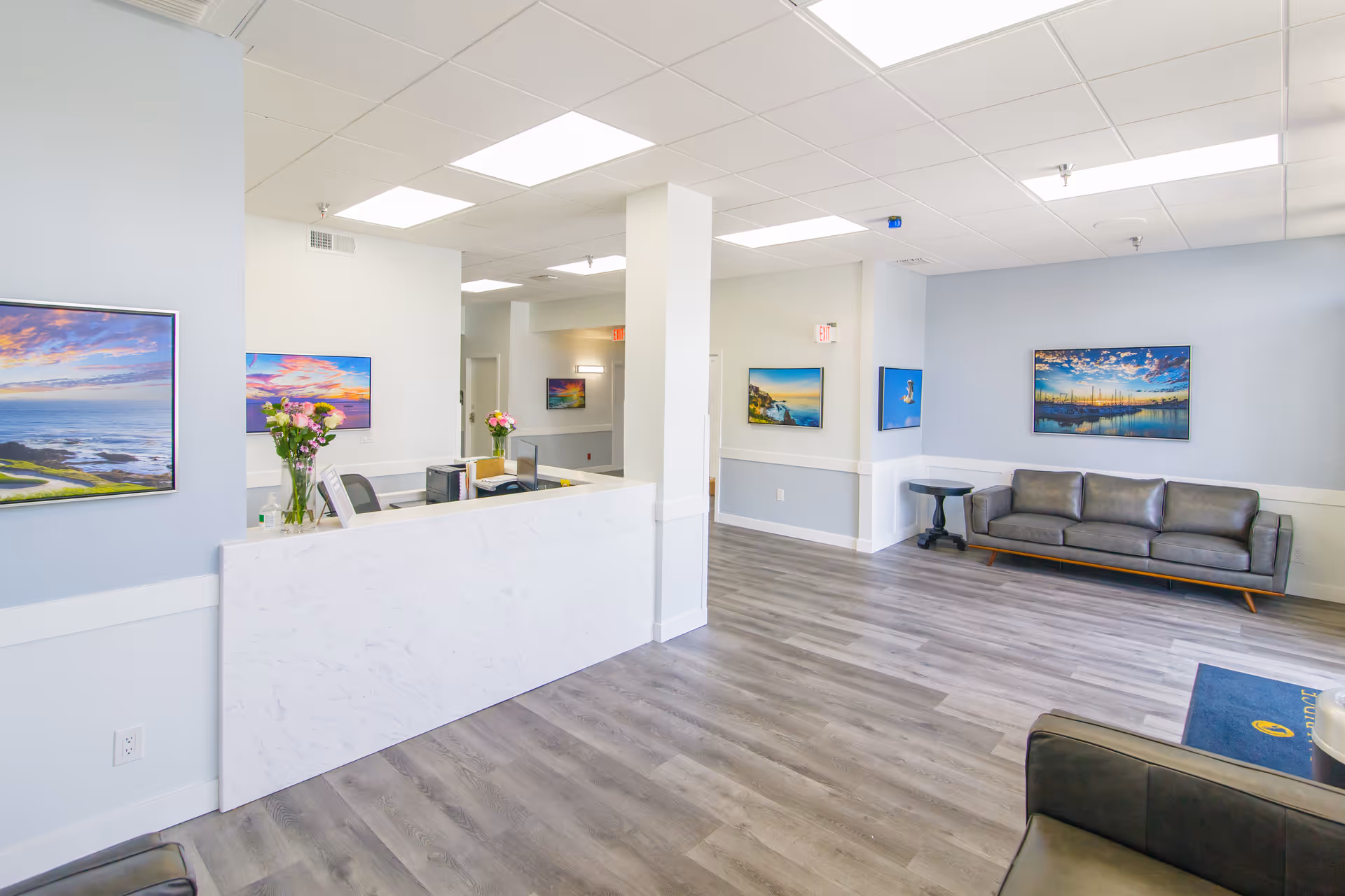 Bright and clean reception area with a white marble desk, a computer, and vases of flowers. The room has light blue walls adorned with colorful landscape paintings, gray wood flooring, and modern gray leather sofas with a small black side table.