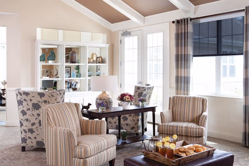 A bright and cozy living room area with two striped armchairs and two floral armchairs around a dark wooden table with a lamp and flower arrangements. A tray with iced tea and pastries is placed on a dark ottoman in the foreground. Large windows with striped curtains allow natural light to fill the room.