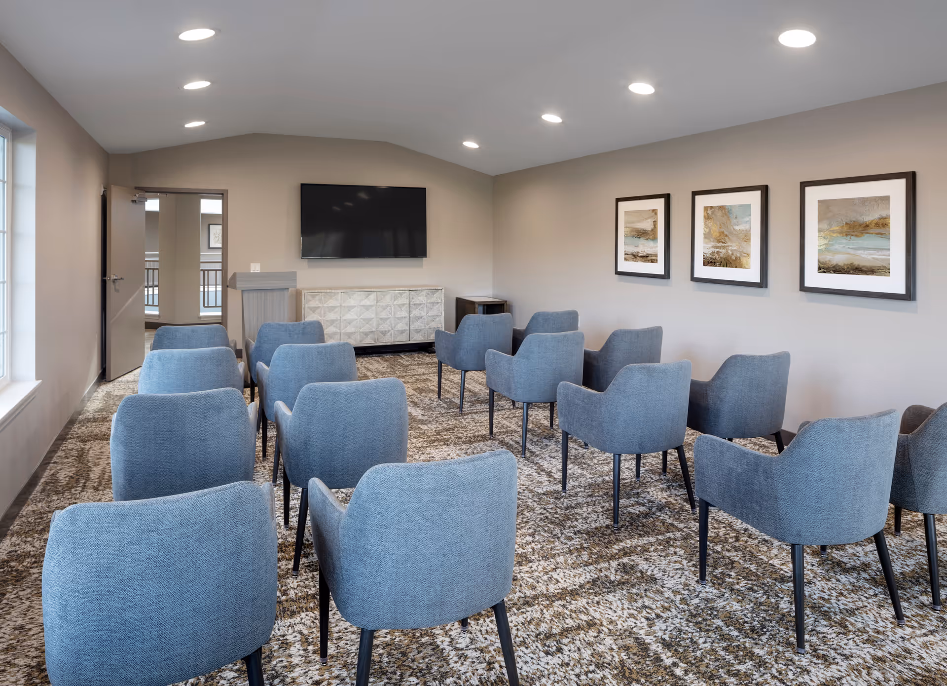 A small conference or meeting room with rows of blue upholstered chairs facing a wall-mounted flat screen TV. The room has beige walls, a patterned carpet, three framed abstract paintings on the right wall, and recessed ceiling lights. There is a podium and a cabinet beneath the TV, and a door on the left side leading to another area.