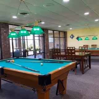Interior view of a recreational room with two pool tables, green hanging lights above them, wooden chairs and tables, and large windows with brick walls in the background.