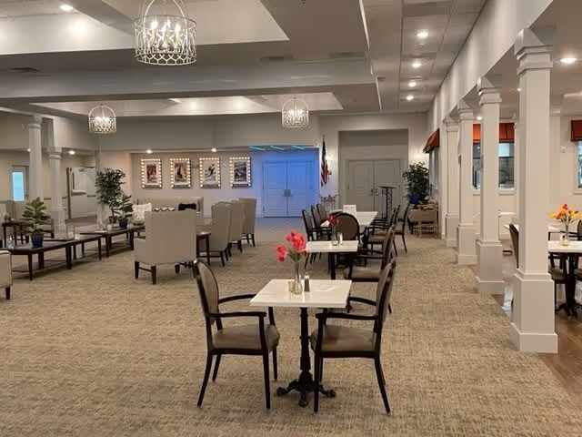 A spacious and well-lit senior living facility common area with multiple tables and chairs arranged for dining or socializing. The room features beige carpeting, white walls with columns, and modern chandeliers hanging from the ceiling. There are plants and framed pictures on the walls, with an American flag near double doors in the background.