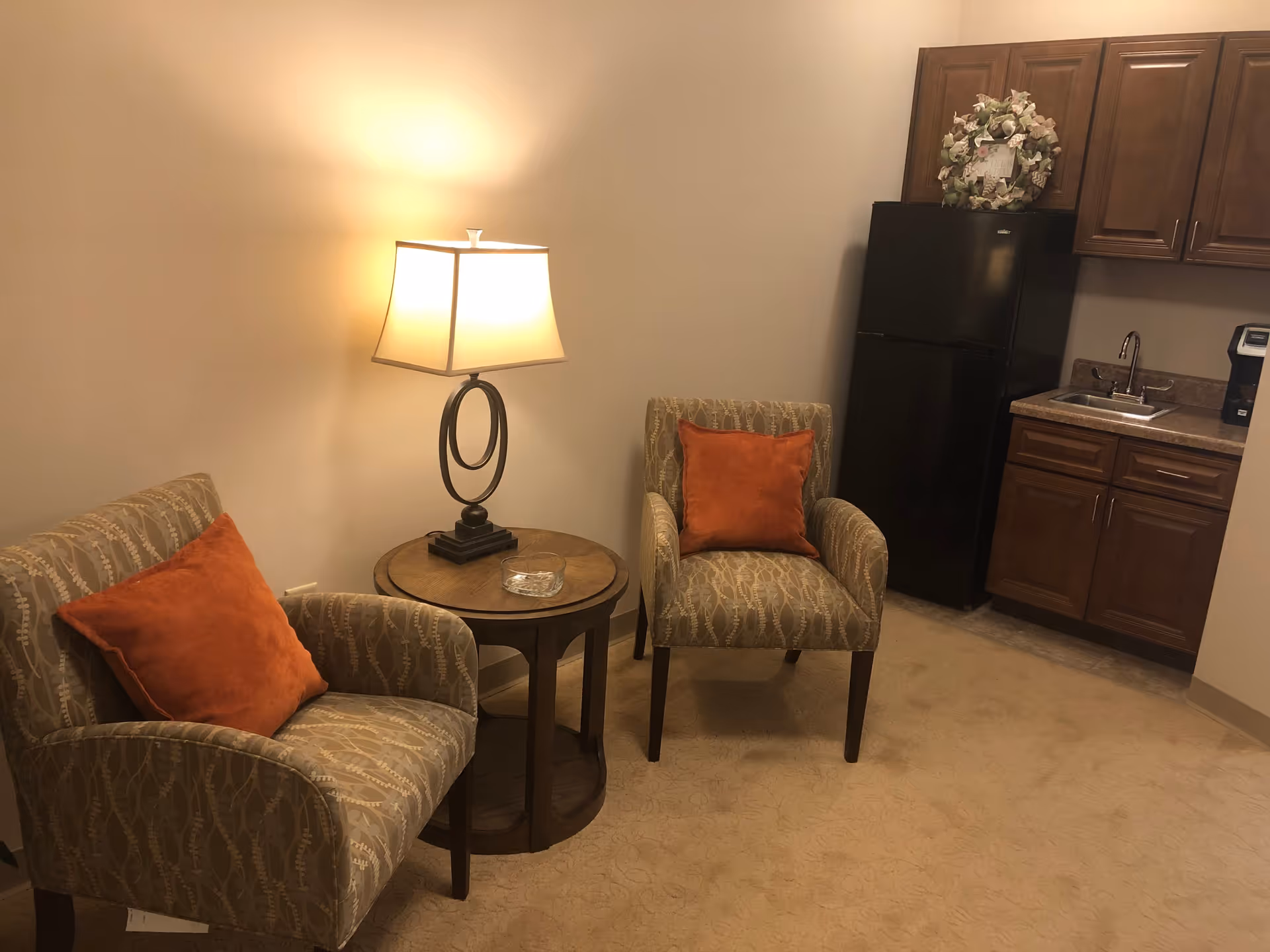 A cozy interior corner featuring two patterned armchairs with orange throw pillows, a round wooden side table with a decorative lamp, and a kitchenette area with wooden cabinets, a black refrigerator, a sink, and a coffee maker.