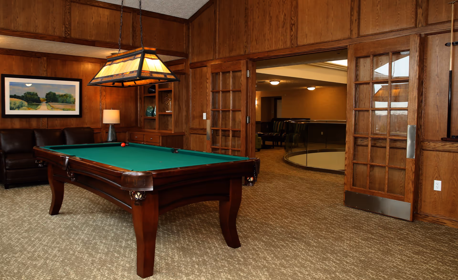Interior room with a green felt pool table in the center, wooden paneled walls, a brown leather couch, a framed landscape painting, and a hanging stained glass light fixture above the pool table. There are double wooden doors with glass panels leading to another room with carpeted flooring and seating.