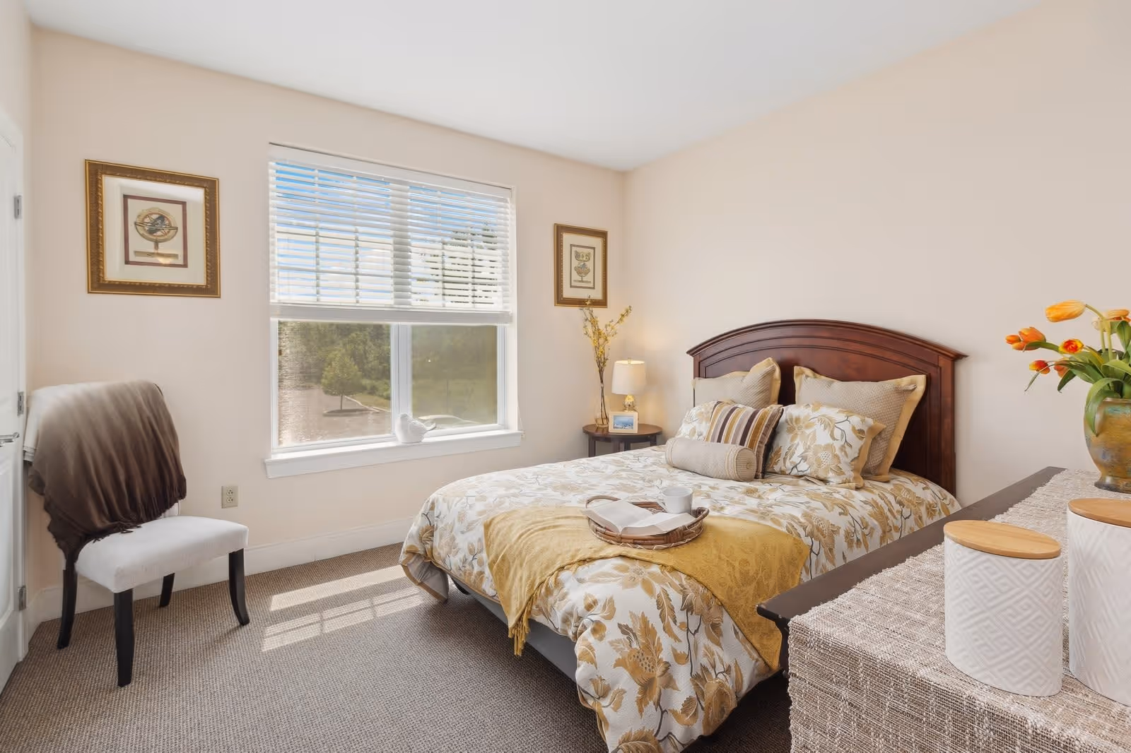 A bright and cozy bedroom with a large window letting in natural light. The room features a bed with floral patterned bedding and multiple pillows, a wooden headboard, a side table with a lamp and decorative items, a chair with a draped blanket, and a dresser with a vase of flowers and decorative containers.